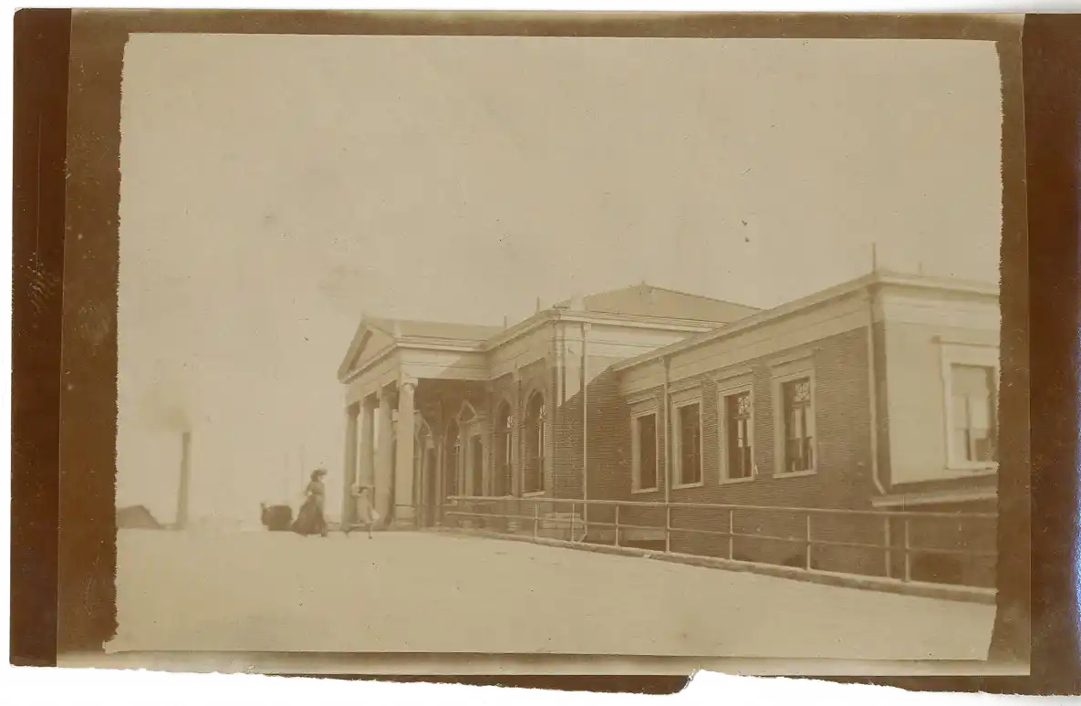 Sepia-toned photograph of a large building with classical architecture, including columns at the entrance, a triangular pediment, and multiple windows along the side. Two people are walking near the entrance, and there is a smokestack visible in the distant left.