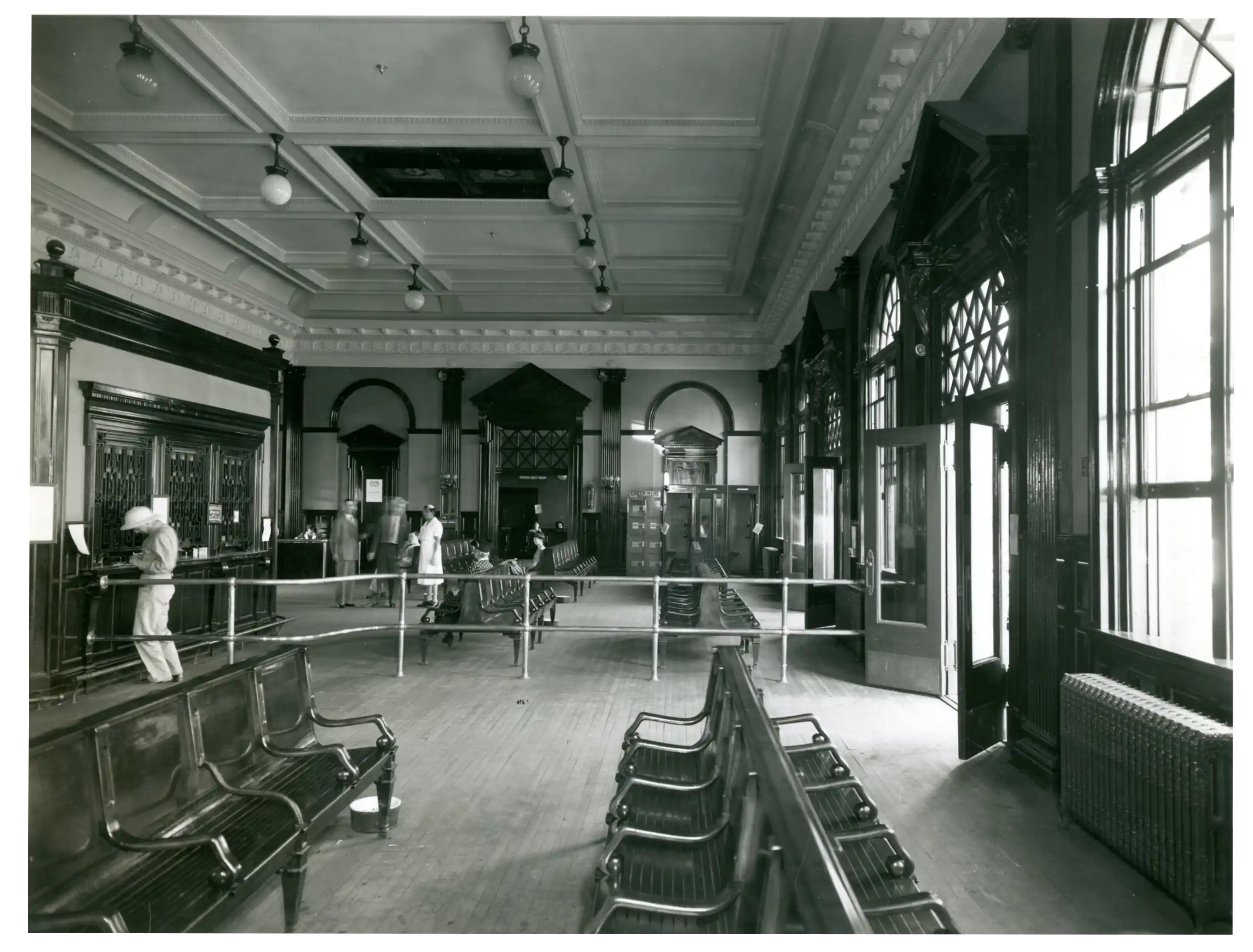 Black and white photo of a large waiting room with wooden floors and rows of empty wooden chairs with metal armrests, high ornate ceiling with hanging light fixtures, and tall arched windows and doors on the right side, few people standing and walking in the distance near a ticket counter on the left.
