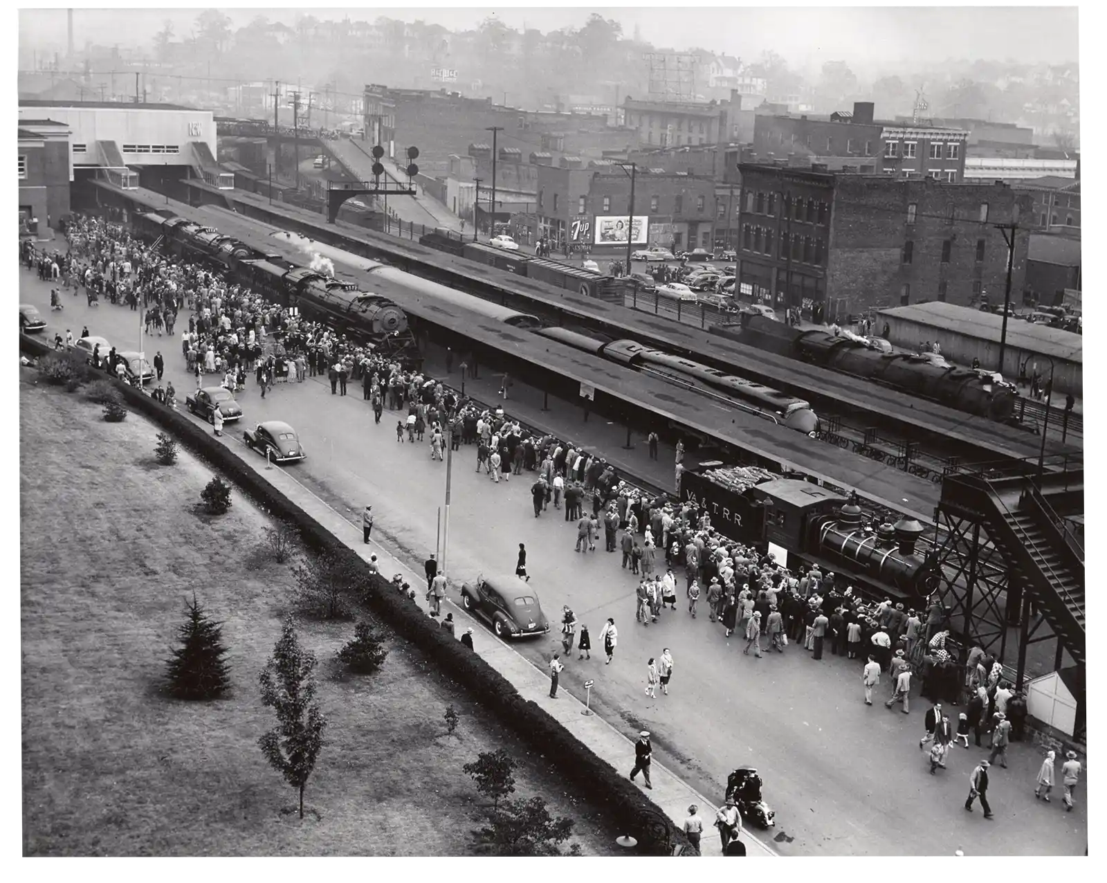 Black and white aerial photo of a busy train station with a crowd of people gathered along the platform next to several steam locomotives labeled W.A.T.R.R. Several vintage cars are parked on the adjacent road, and old buildings and a hilly cityscape are visible.