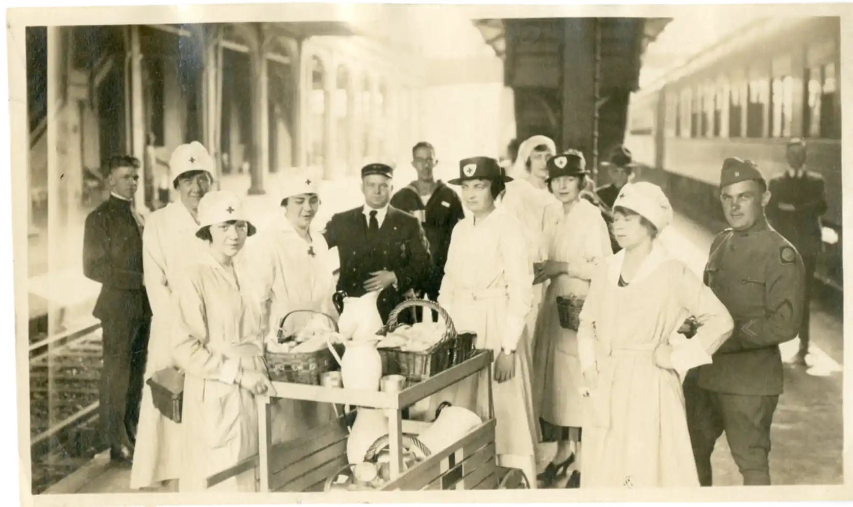 Black and white photo depicting a group of nurses and soldiers standing around a cart with baskets and pitchers on a train station platform with a train visible on the right side.