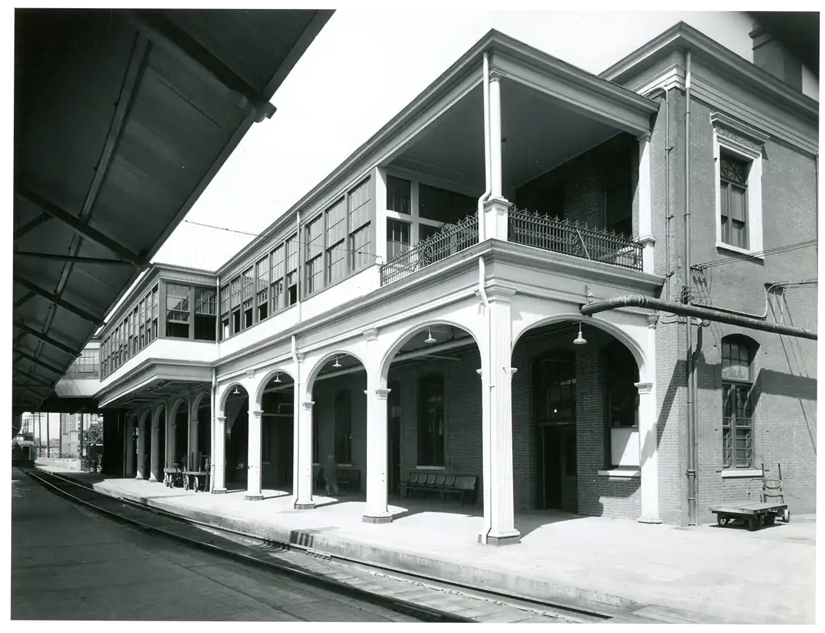 Black and white photo of a historic two-story brick train station building with a covered platform and multiple arched openings supported by columns.