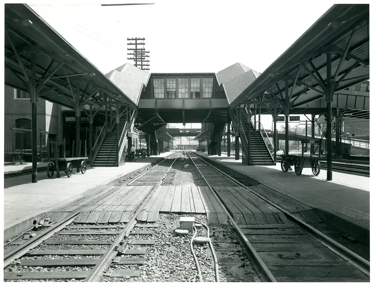 Historic black and white photo of a railway station showing train tracks in the center flanked by covered platforms with staircases leading to an overhead pedestrian bridge connecting both sides.