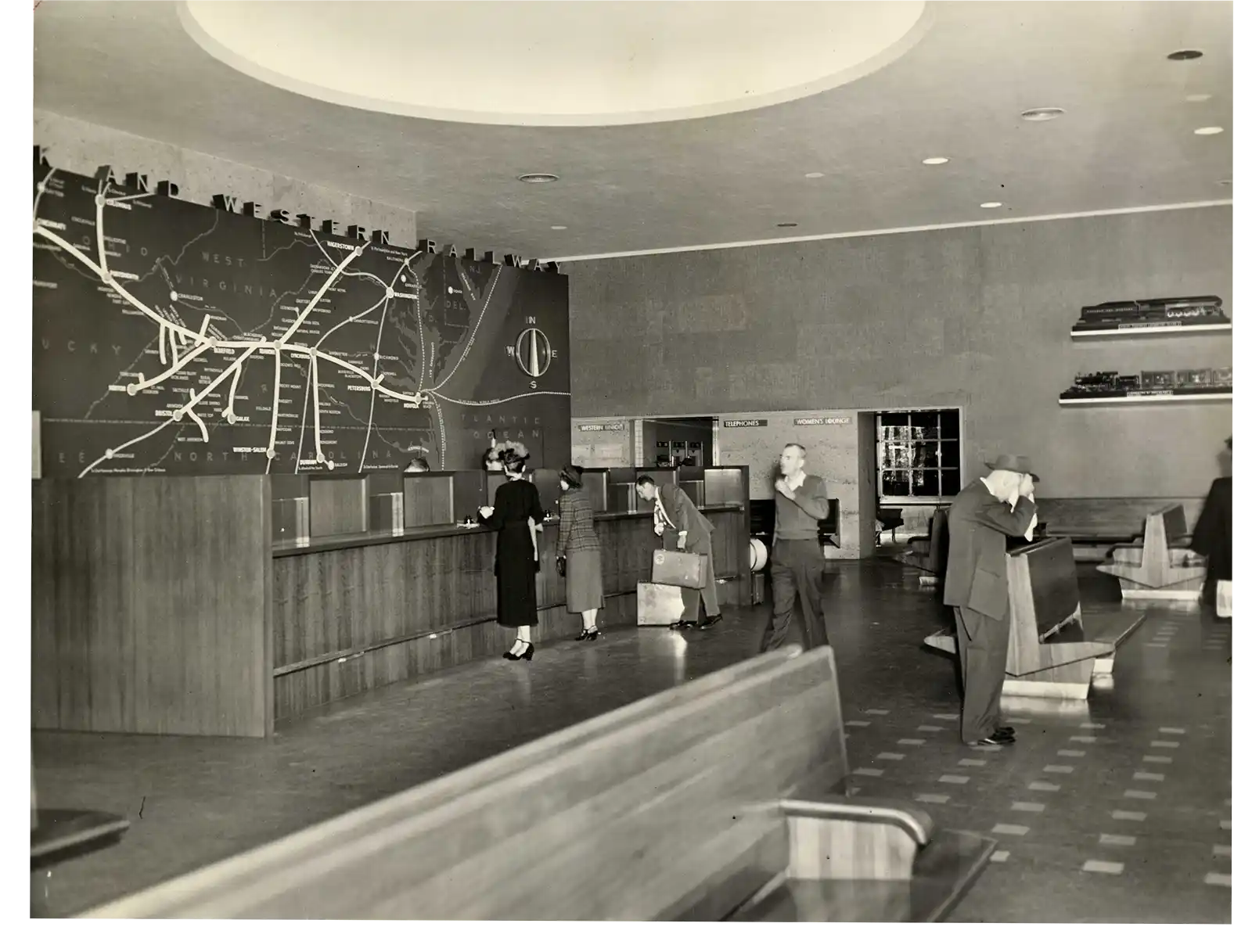 Black and white photo of a railway station ticket counter with a large route map on the wall, showing lines and station names. Several people stand at the counter or nearby, some holding luggage, engaging in typical travel activities. The room features benches, a high ceiling with a circular light fixture, and shelves mounted on the far wall holding model trains or similar objects.