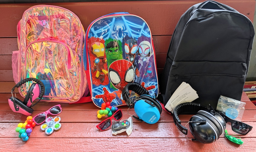 Three backpacks on a wooden bench: a shiny pink one, a blue one with Marvel superheroes, and a plain black one, each accompanied by sensory items including colorful fidget toys, sunglasses, and protective earmuffs in pink, blue, and black.