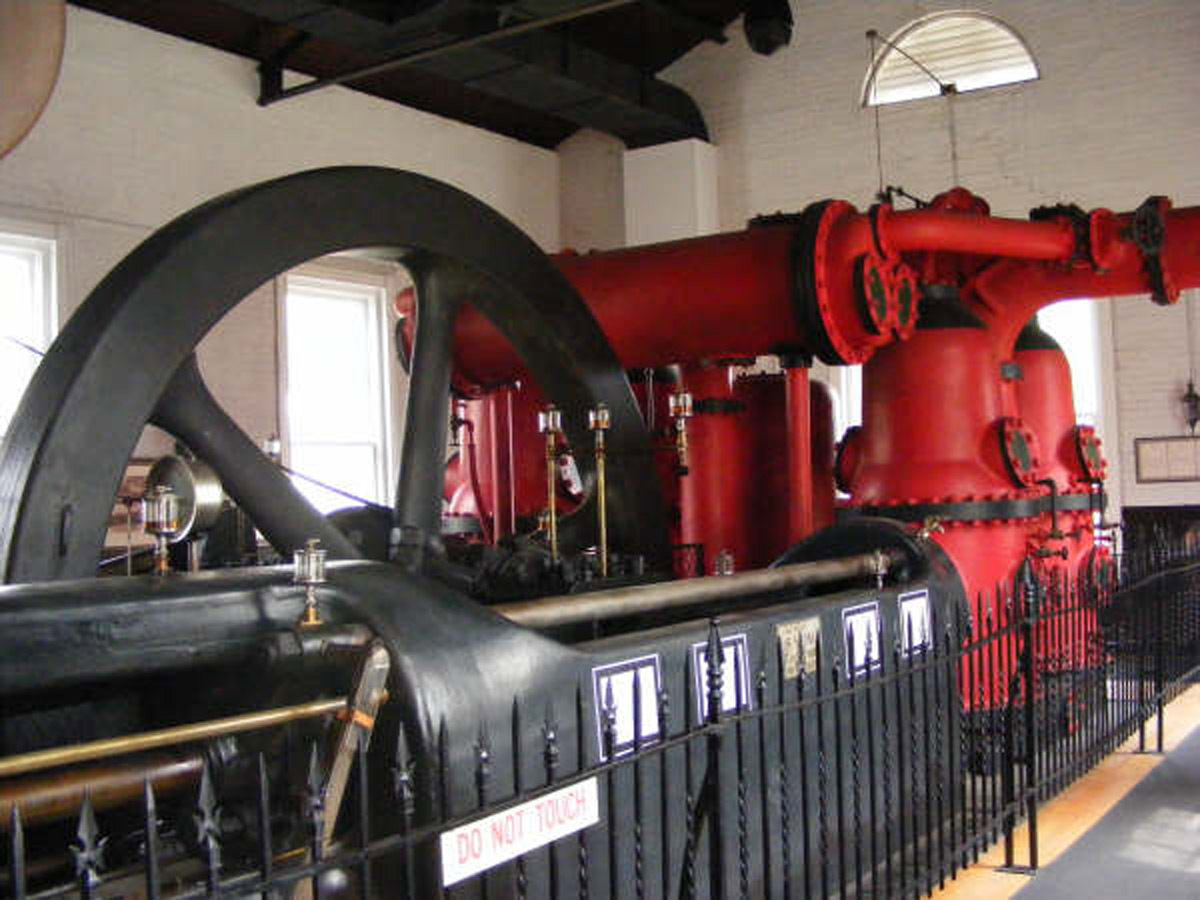 Large industrial steam pump with a prominent black flywheel and red piping inside a building, behind a black metal fence with a 'DO NOT TOUCH' sign.