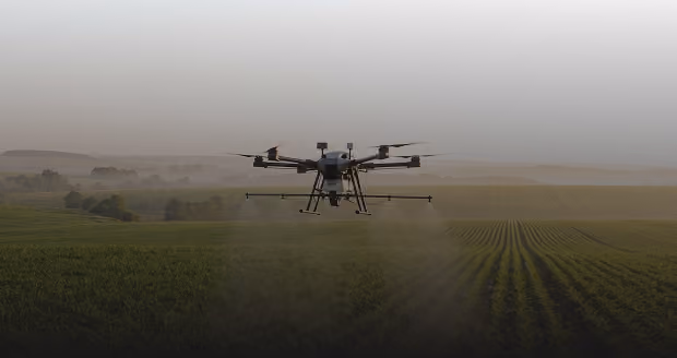 Agricultural drone spraying crops over a large green field at dusk with soft rolling hills and trees in the background.