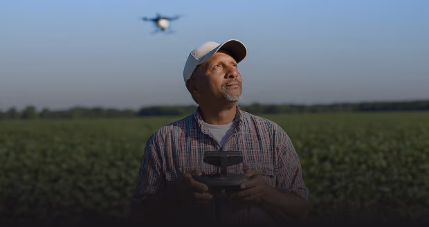 Man wearing a cap and plaid shirt operating a drone controller in a green field with the drone flying in the clear blue sky.