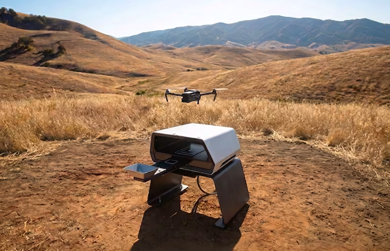 A drone hovers above a modern drone docking station with an open compartment in a dry, grassy field surrounded by rolling hills under a clear sky.