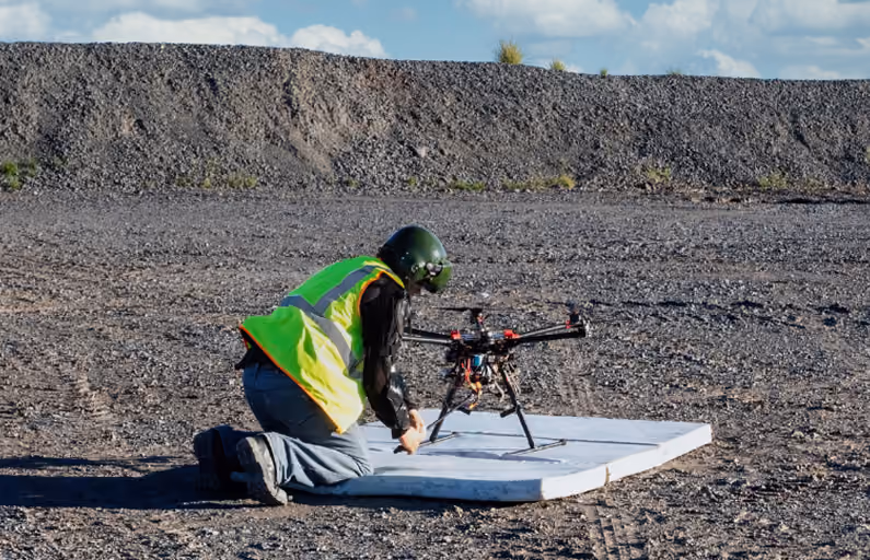 Person wearing a green helmet and neon yellow safety vest kneeling on gravel while preparing a multi-rotor drone on a foam launch pad outdoors under a blue sky.