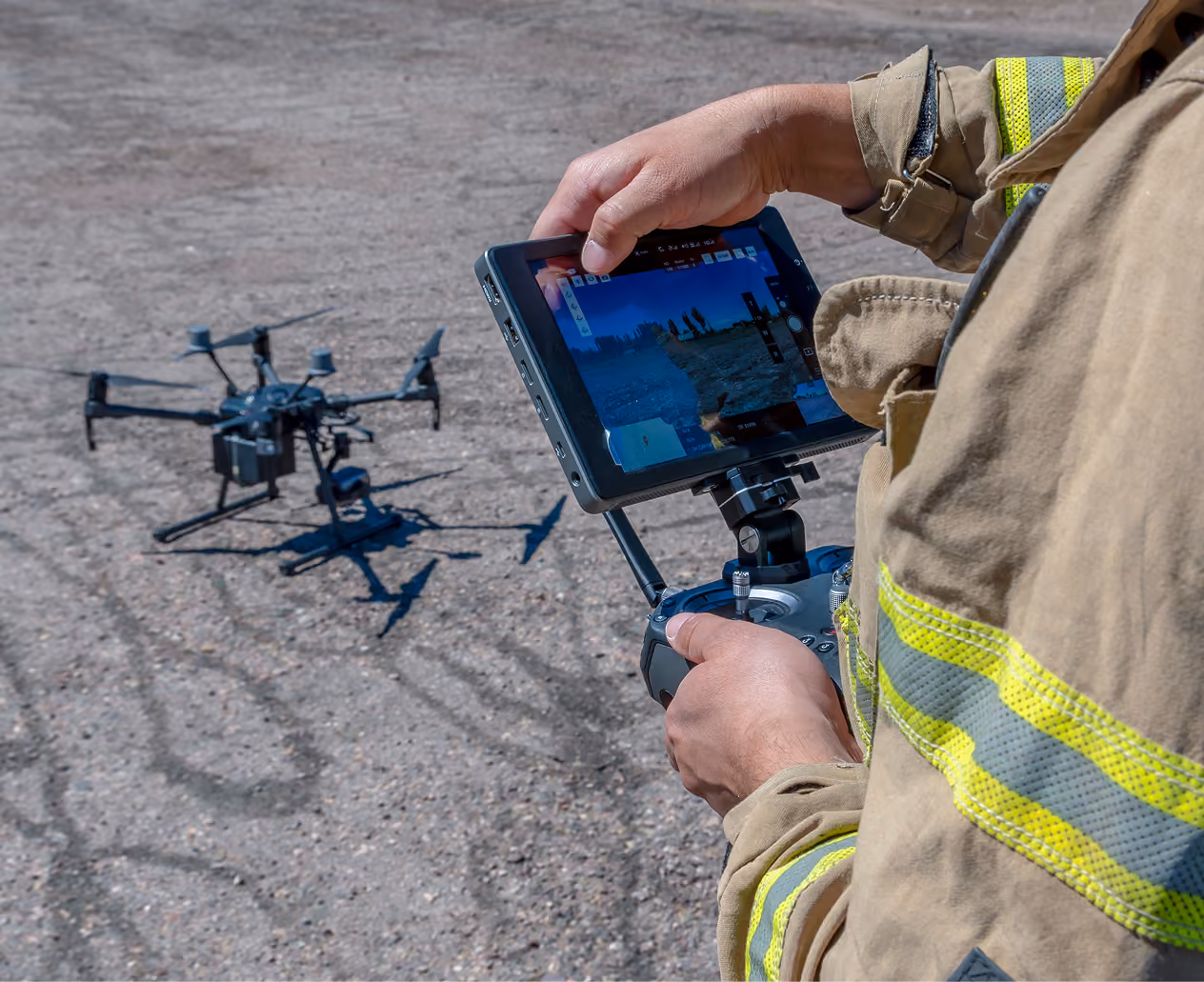 Person in a tan jacket with yellow reflective stripes operating a drone using a remote control with attached screen showing live drone footage of an outdoor dirt area.