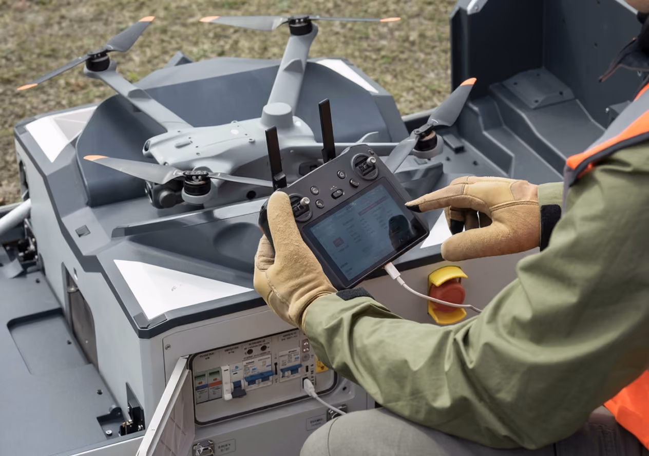 Person wearing gloves and a green jacket operates a remote control device with a screen, connected by a cable to a large drone with four propellers resting on a gray platform.