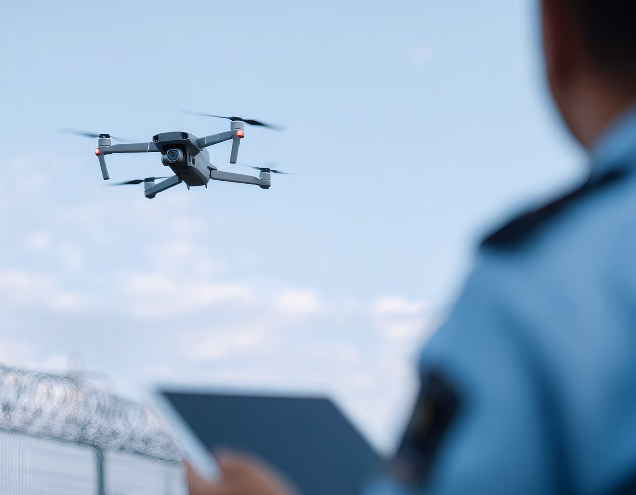 Drone hovering in the air controlled by a person holding a tablet, with a barbed-wire fence visible in the background.