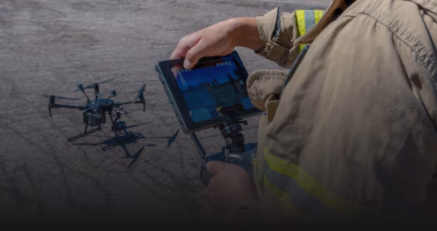 Person in beige jacket with reflective safety stripes operating a drone controller displaying drone flight footage while the drone stands on the ground.