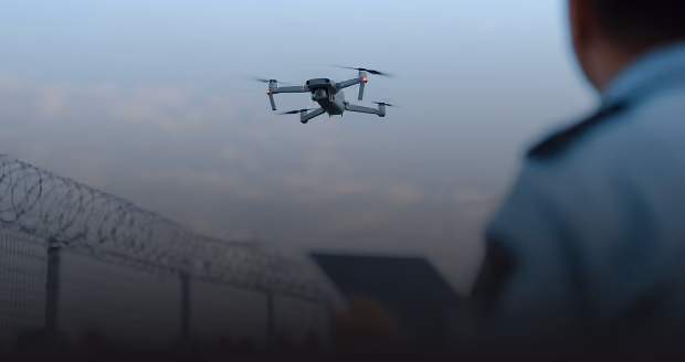 A drone flying in the sky near a barbed wire fence, with a person in uniform holding a tablet, monitoring the drone.