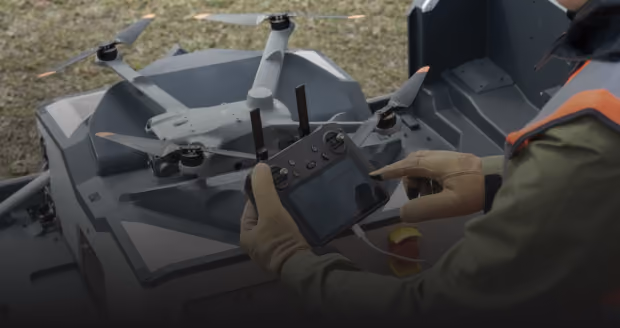 Person wearing gloves holding a drone remote controller, operating a gray quadcopter drone on a gray surface outdoors.