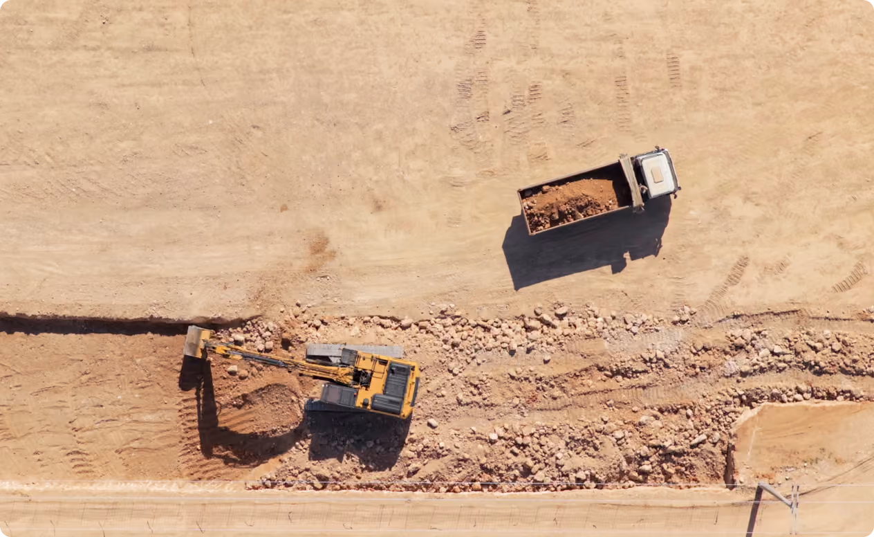 Aerial view of a construction site with a yellow excavator digging in dirt and a white dump truck nearby carrying a load of soil and rocks.