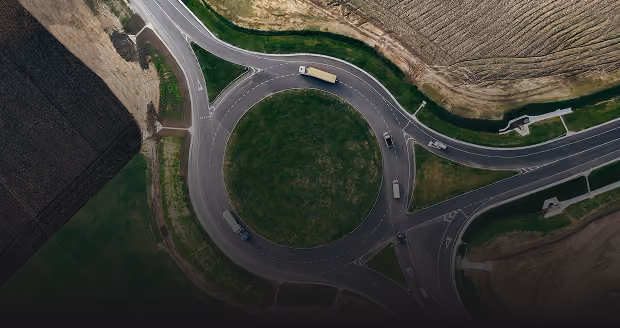 Aerial view of a roundabout with a grassy center surrounded by multiple lanes of traffic, including several trucks and cars, with agricultural fields adjacent to the road.