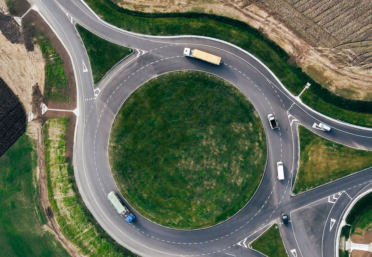Aerial view of a circular roundabout with green grass in the center, surrounded by multiple vehicles including trucks and cars on the asphalt road.