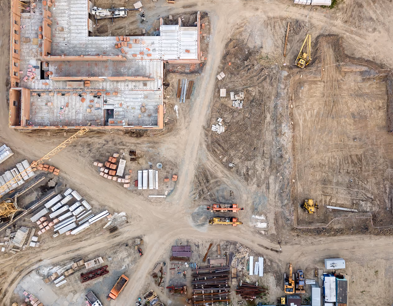Aerial view of a construction site with a partially built concrete structure, construction materials, heavy machinery, and dirt roads forming a cross shape.