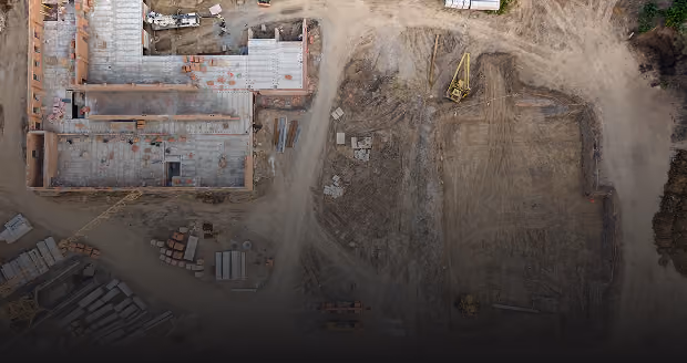 Aerial view of a construction site with a partially built brick and concrete structure on the left, construction materials, and earth-moving equipment on cleared dirt areas on the right.