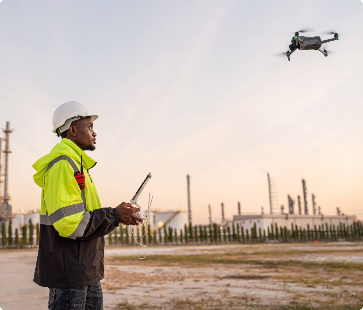 Man wearing a white hard hat and neon green safety jacket operating a flying drone with a remote controller at an industrial site during dusk.