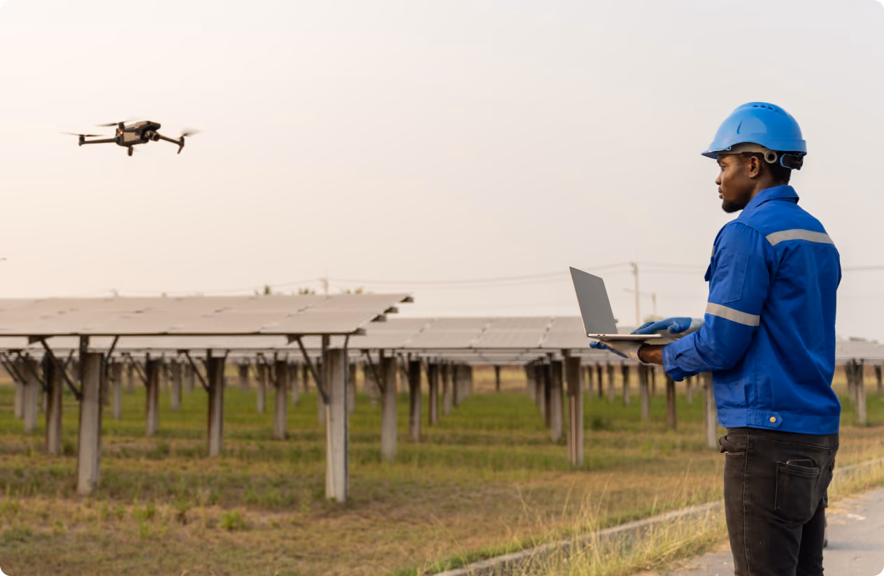 Technician in blue safety gear operates a drone using a laptop near a solar panel farm during daylight.