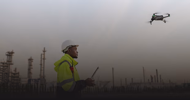 A worker wearing a white hard hat and high-visibility jacket operates a drone with an industrial refinery in the background at dusk or dawn.