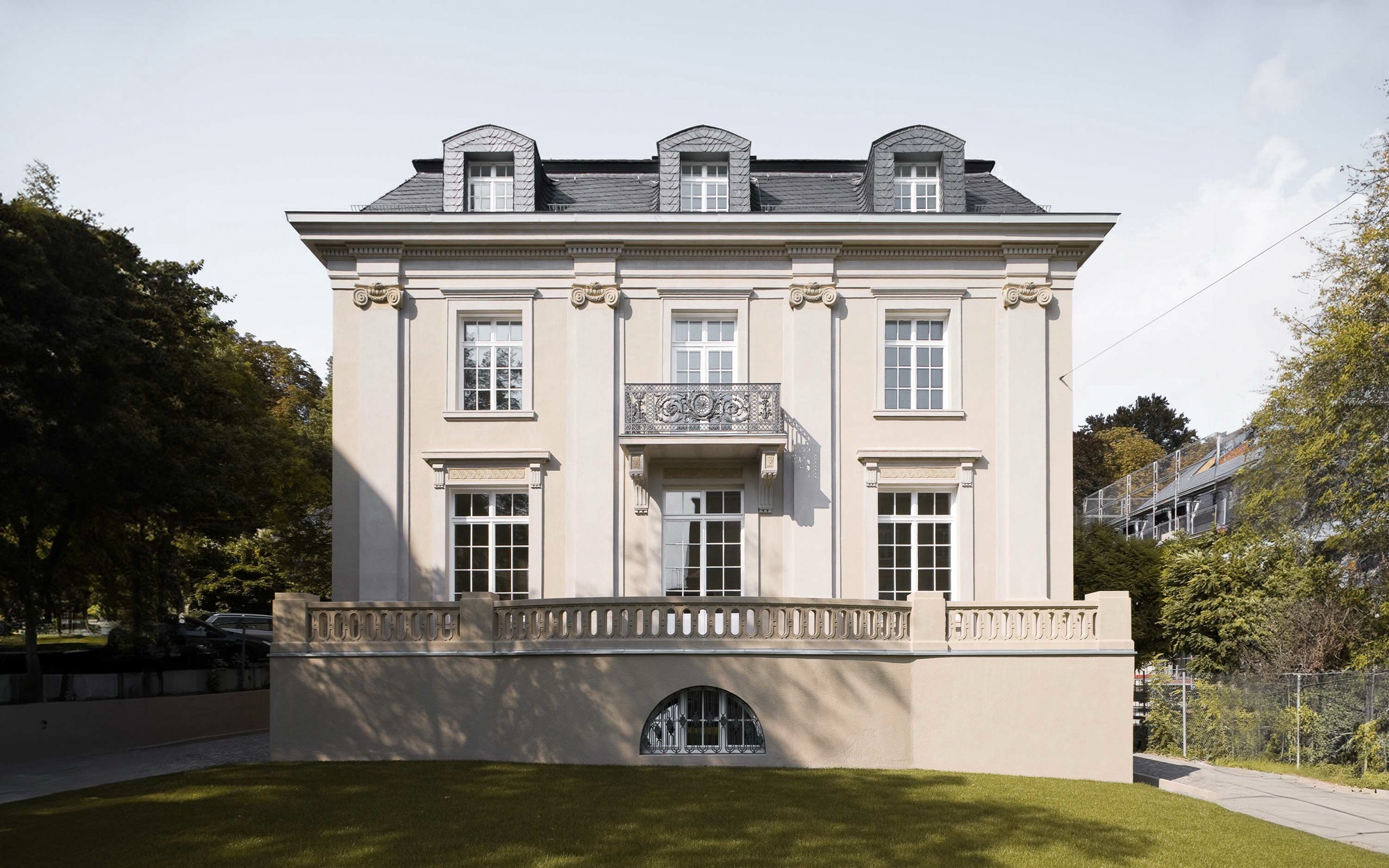 Front view of a classic beige mansion with tall windows, decorative balcony, and dormer windows on roof.