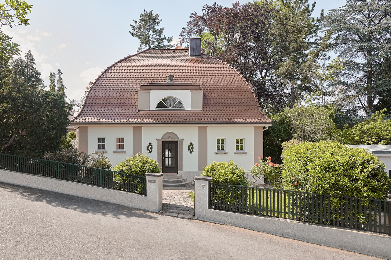 Front view of a classic beige mansion with tall windows, decorative balcony, and dormer windows on roof.