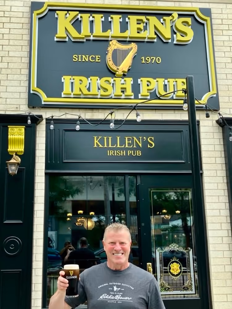 Smiling man holding a pint of Guinness in front of Killen’s Irish Pub entrance and sign.