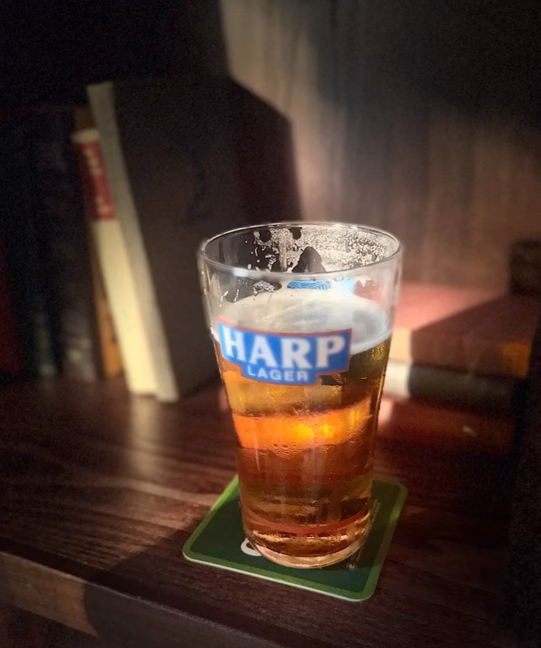 Glass of Harp Lager beer on a wooden table with books in soft focus background.