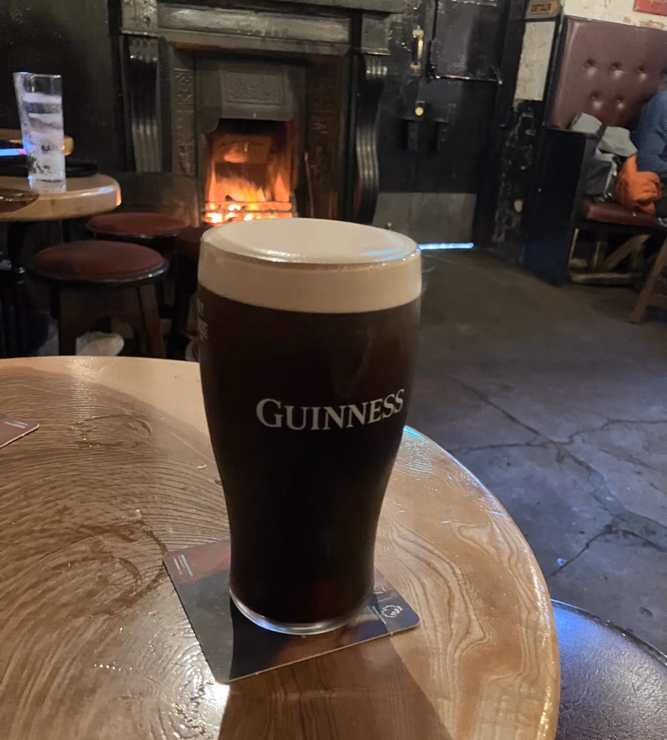 Pint of Guinness stout on a wooden table in a pub with a lit fireplace in the background.