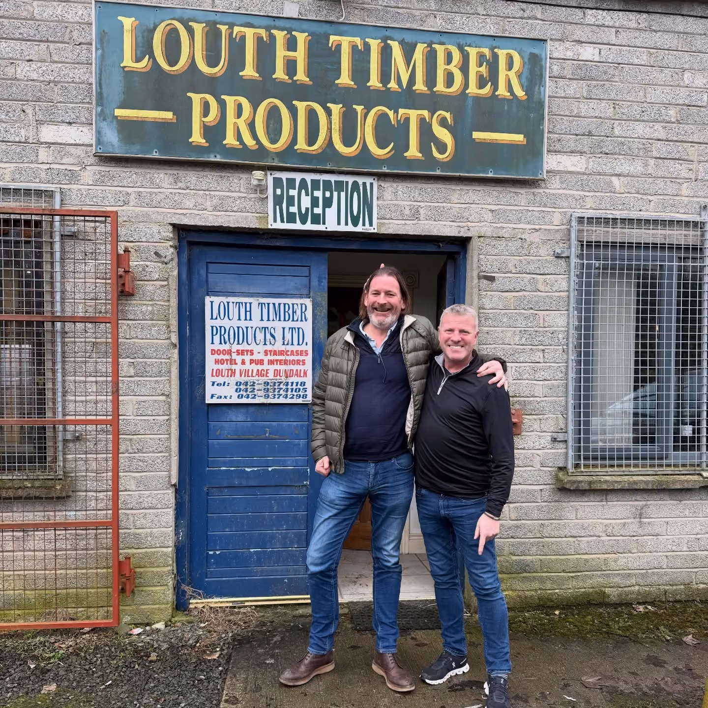 Two smiling men standing arm in arm outside Louth Timber Products shop entrance with blue door and signs.