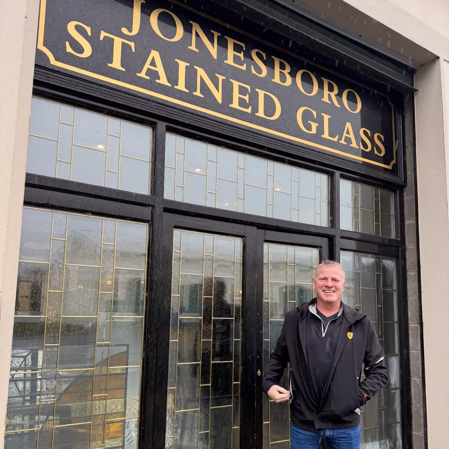 Smiling man in black jacket standing in front of a door with stained glass and a Jonesboro Stained Glass sign.