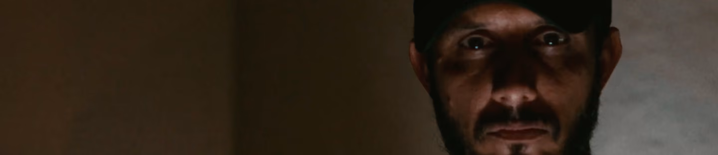 Hombre con barba y gorra mirando fijamente en habitación con luz tenue.