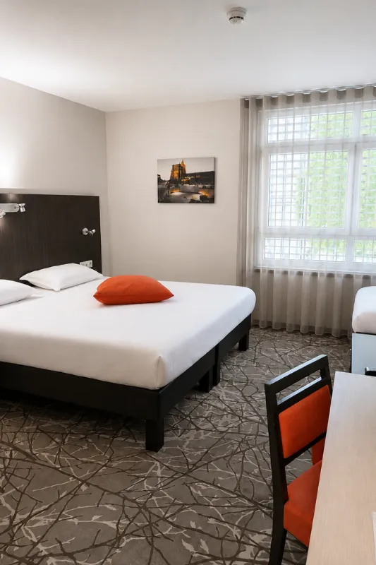 Chambre lumineuse avec lit double, coussin orange, bureau et grande fenêtre voilée