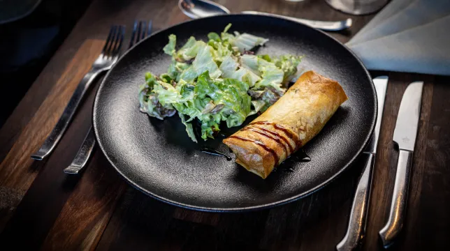 Pastilla croustillante nappée de sauce balsamique, servie avec une salade verte dans une assiette noire sur une table en bois dressée