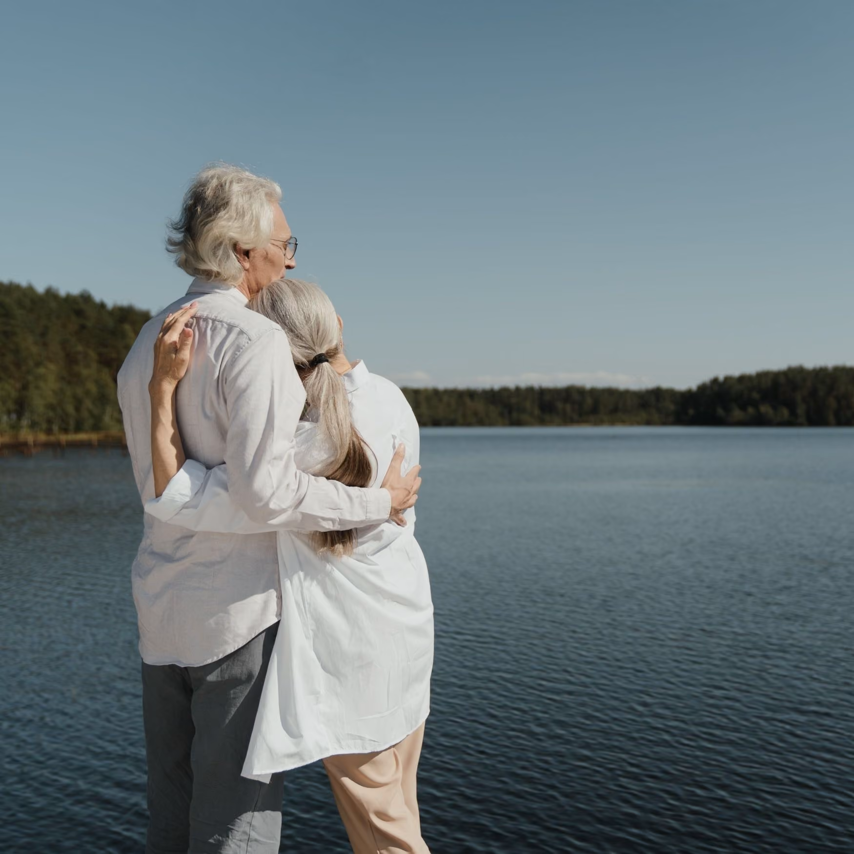 An elderly couple wearing light clothes hugging and looking at a lake under clear blue sky.