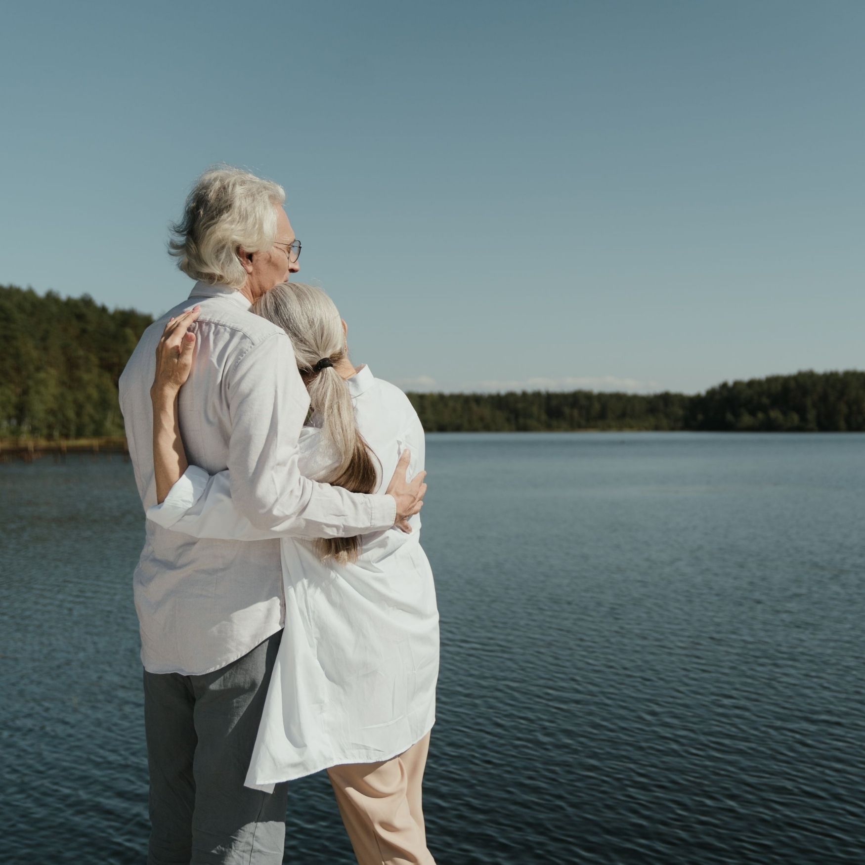 An elderly couple wearing light clothes hugging and looking at a lake under clear blue sky.