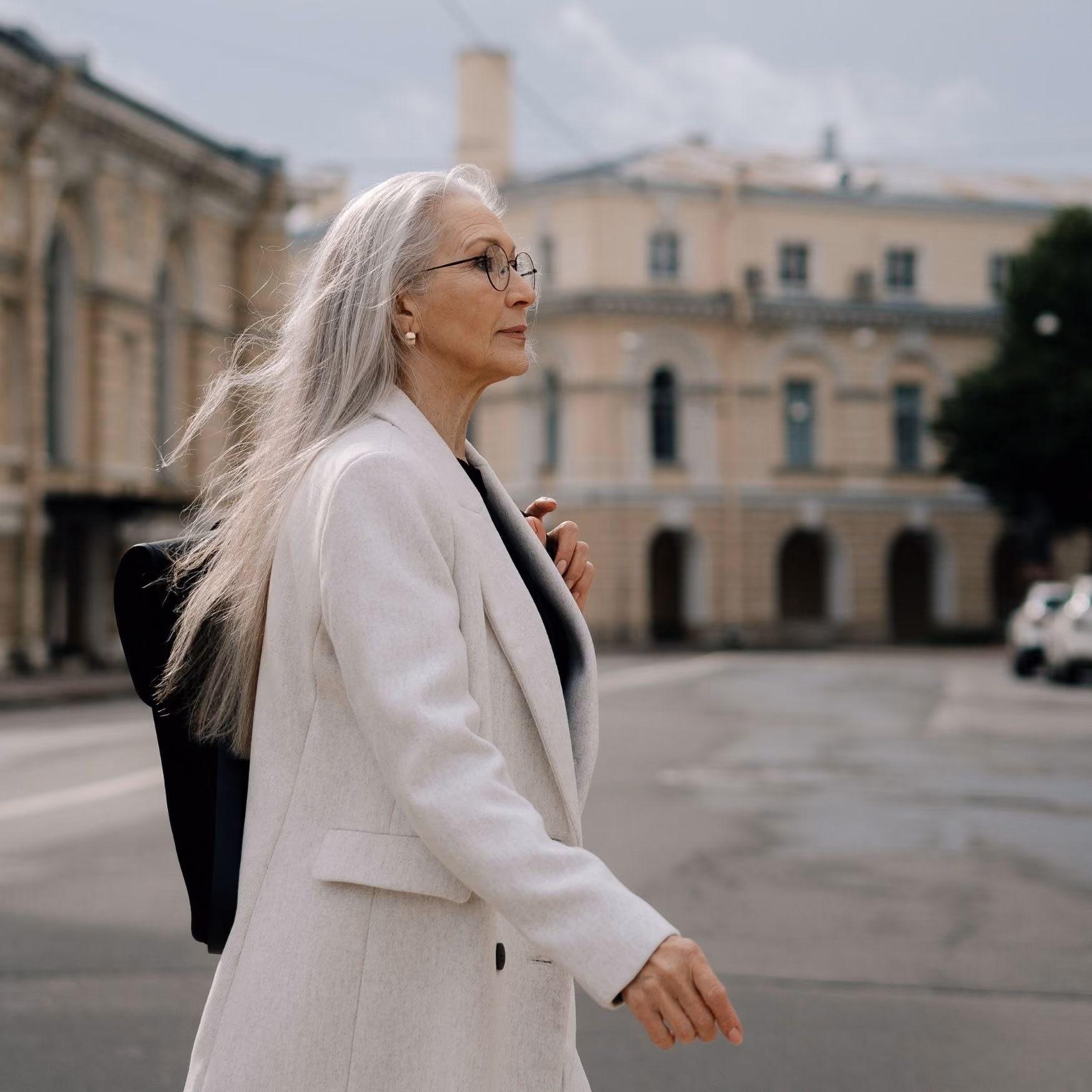 Elderly woman with long gray hair and glasses walking outdoors wearing a light-colored coat.