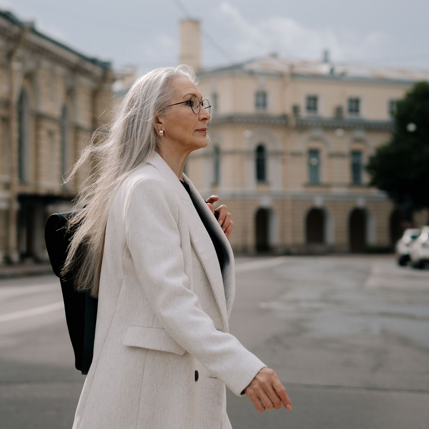 Elderly woman with long gray hair and glasses walking outdoors wearing a light-colored coat.