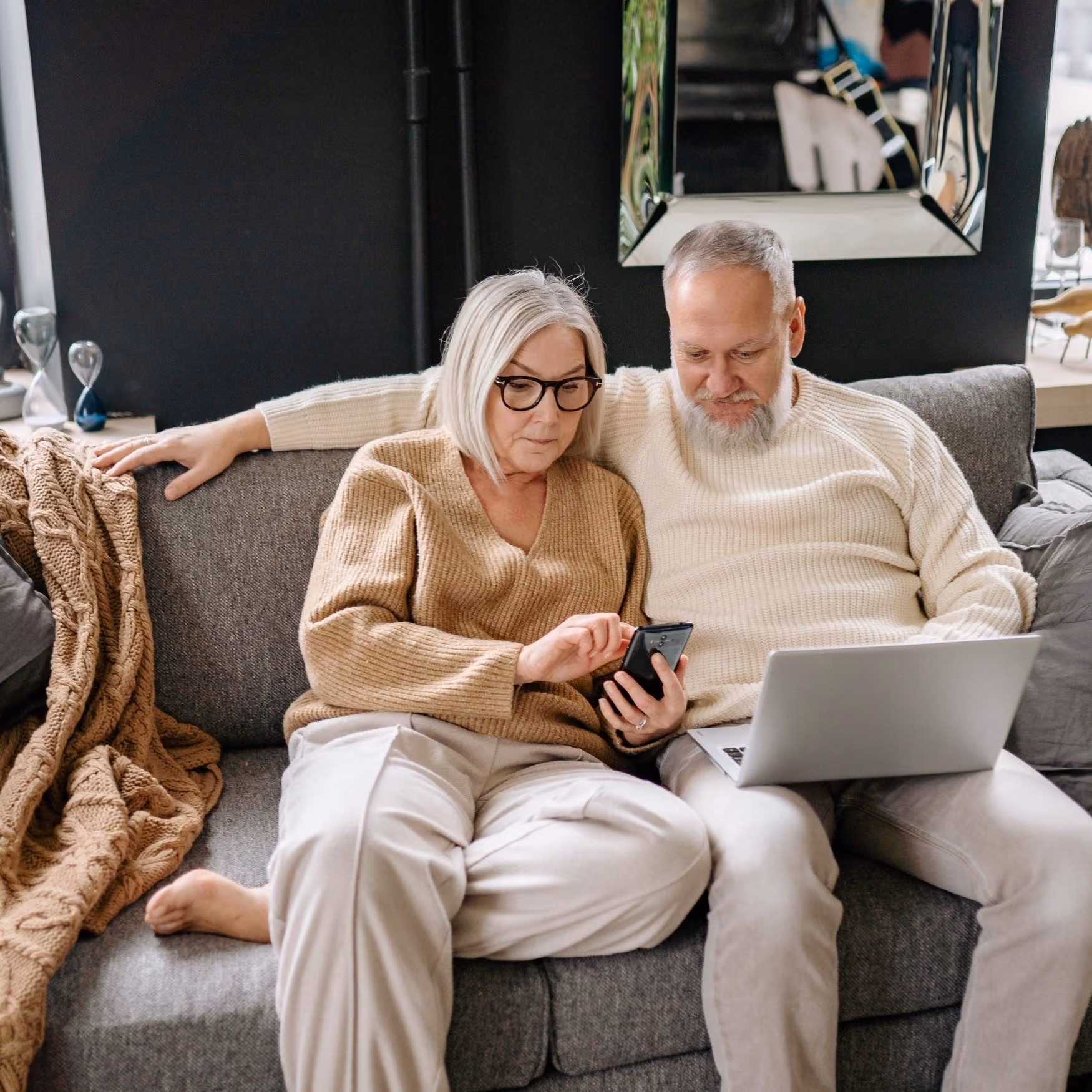Elderly couple sitting on couch, woman using smartphone and man holding laptop, cozy living room setting.