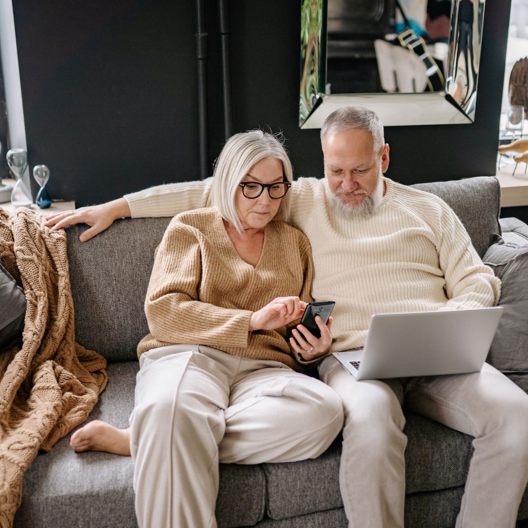Elderly couple sitting on couch, woman using smartphone and man holding laptop, cozy living room setting.