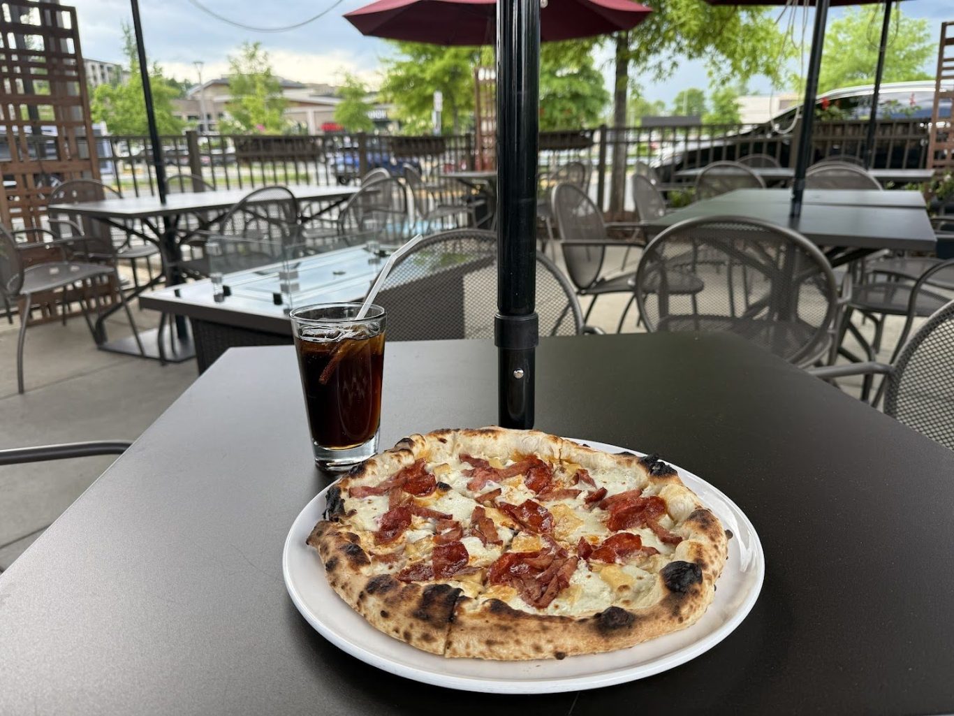 Pepperoni pizza on a white plate with a glass of cola and straw on an outdoor restaurant table.
