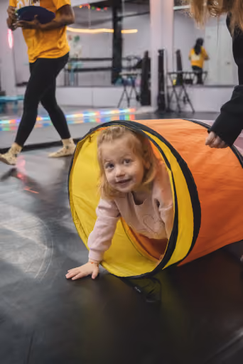 A young dancer smiling from inside a play tunnel during a preschool class at Revolution Dance Academy