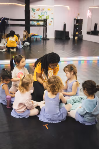 An RDA instructor kneeling with a circle of young dancers on the studio floor