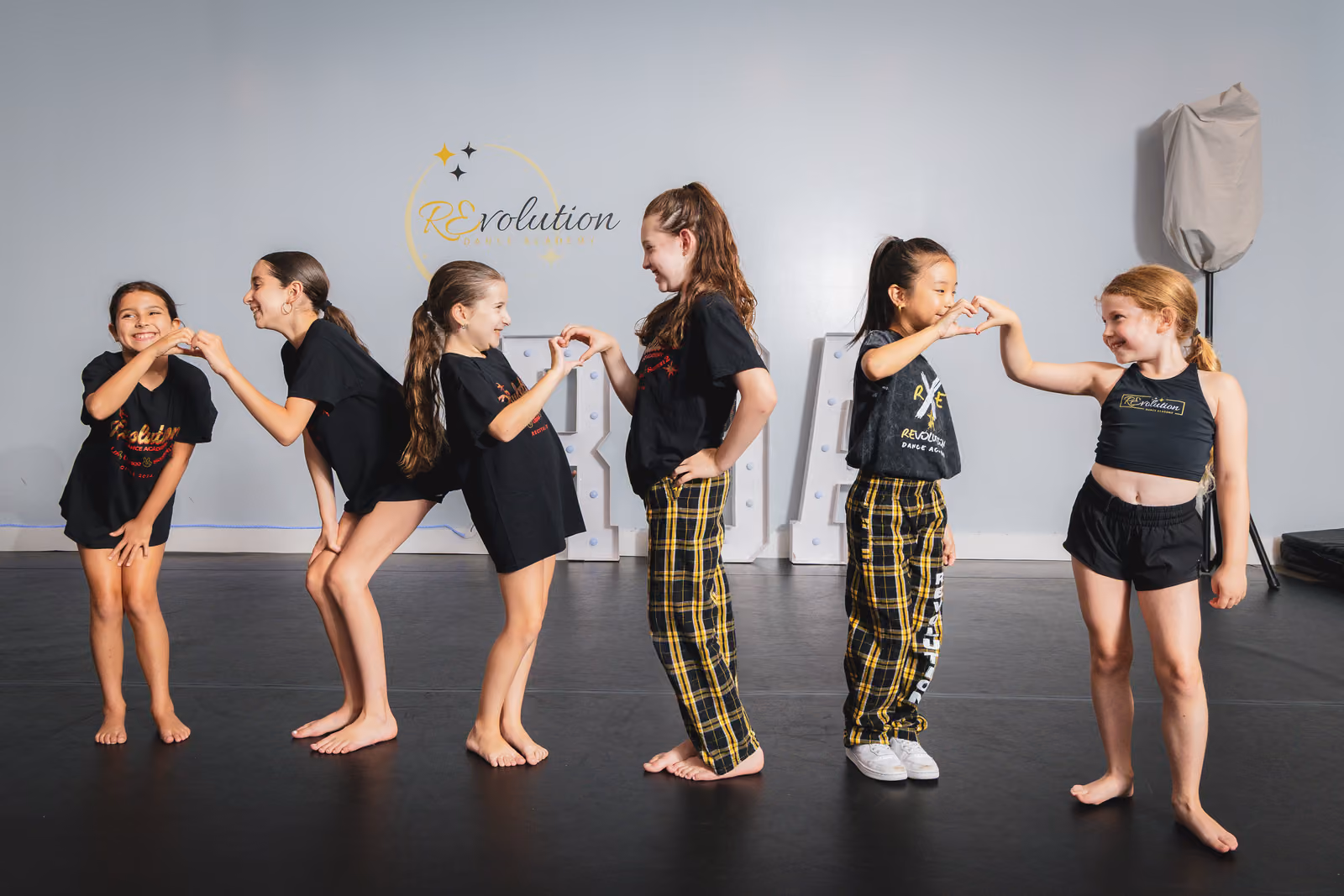Six girls in dance wear form heart shapes with their hands inside a dance studio with REvolution logo.