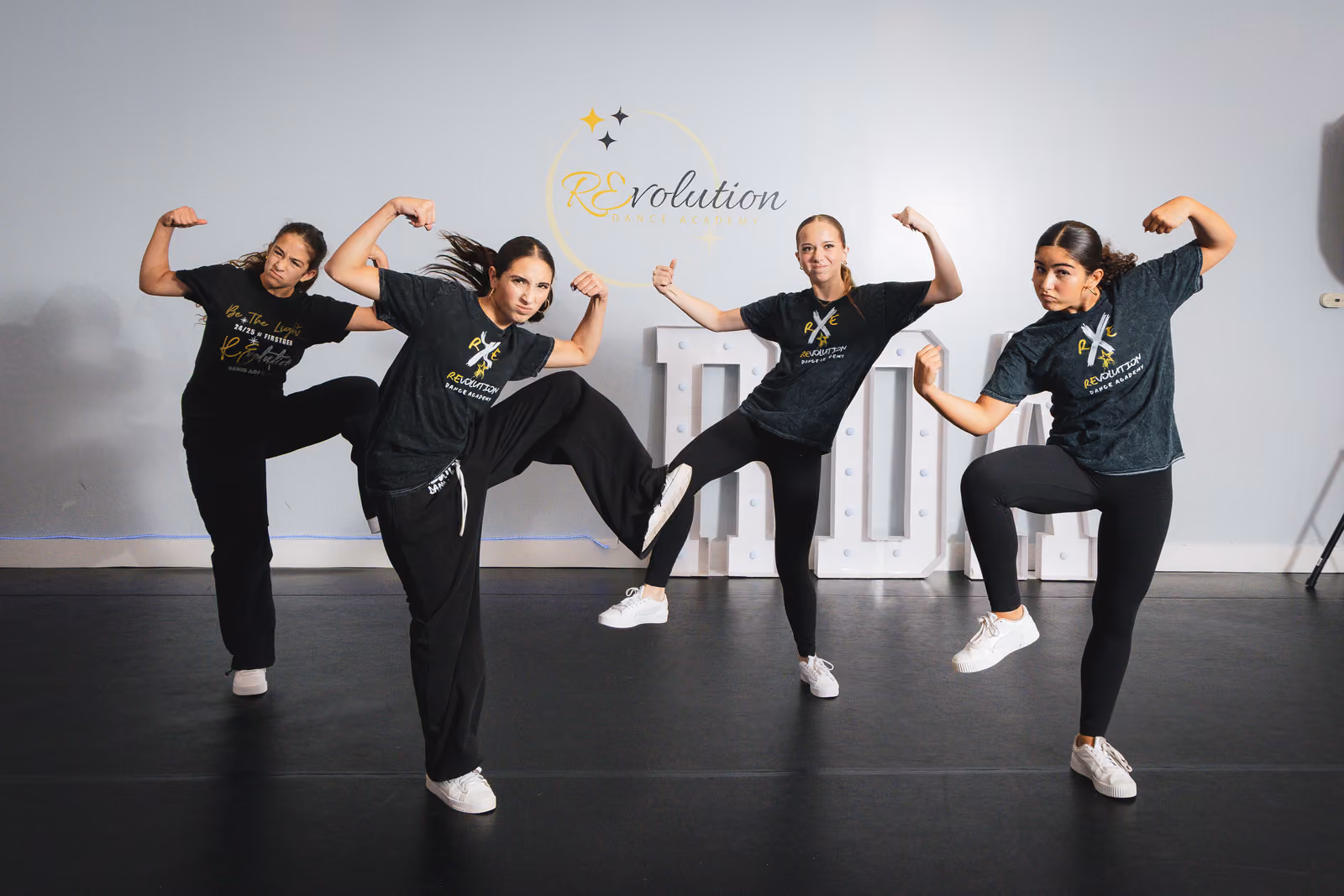 Four young women in black dancewear flex muscles and raise legs in a hip-hop studio with Revolution Dance logo.