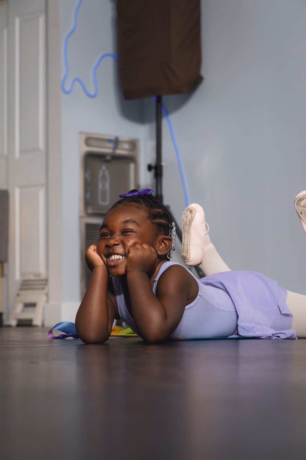 Smiling young girl in purple ballet outfit lying on floor with hands on cheeks and feet raised.