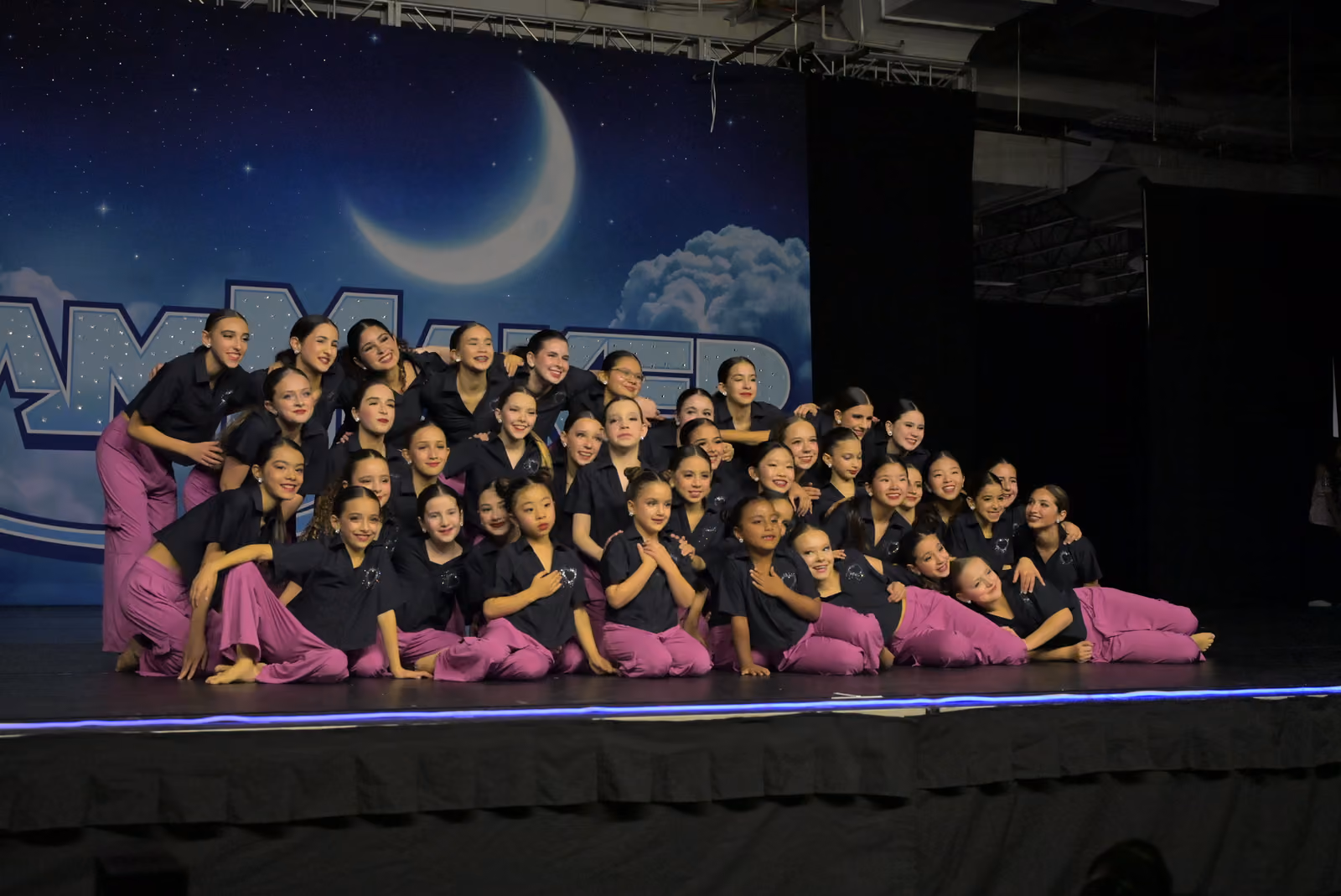 Large group of young dancers in black shirts and purple pants posing on stage under a moon and stars backdrop.
